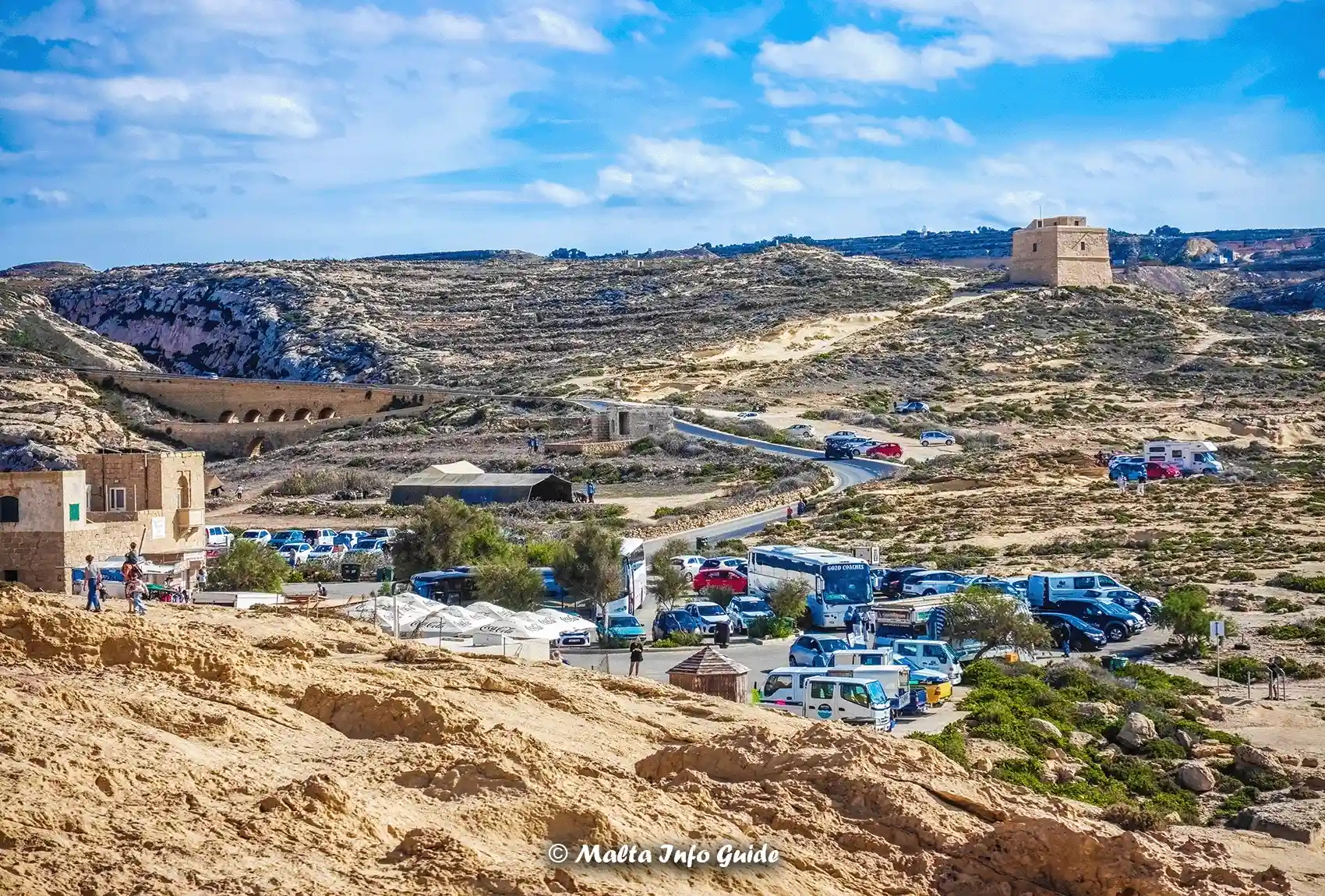 Scenic view of Dwejra Bay with parked vehicles and historical structures.