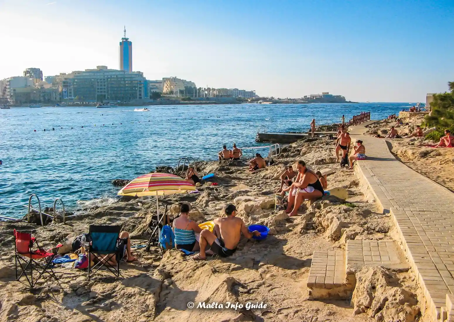 Swimmers relax beside ladders on Exiles rocky Sliema shore.