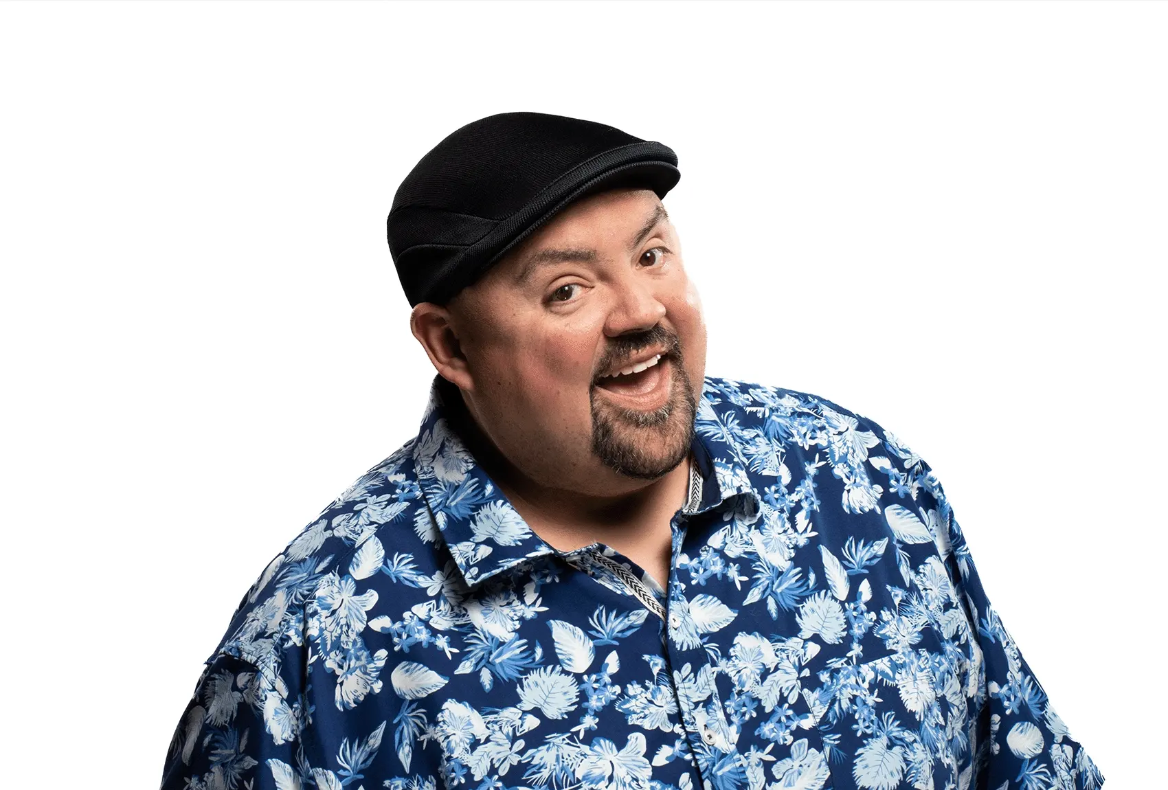 Gabriel Iglesias smiling in a blue floral shirt and black cap against a white background.