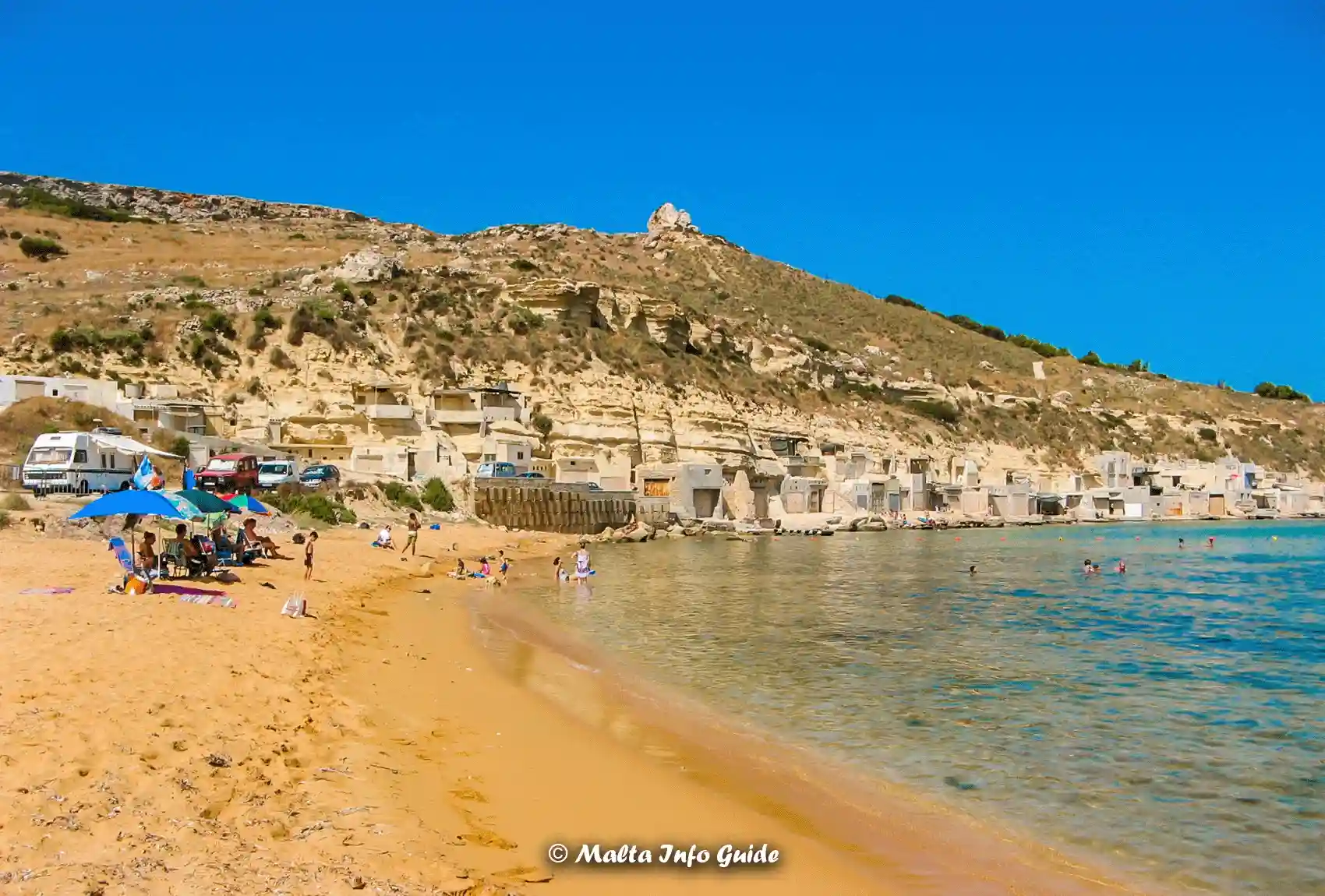 Sandy shoreline and boathouses beside calm Gnejna Bay water.