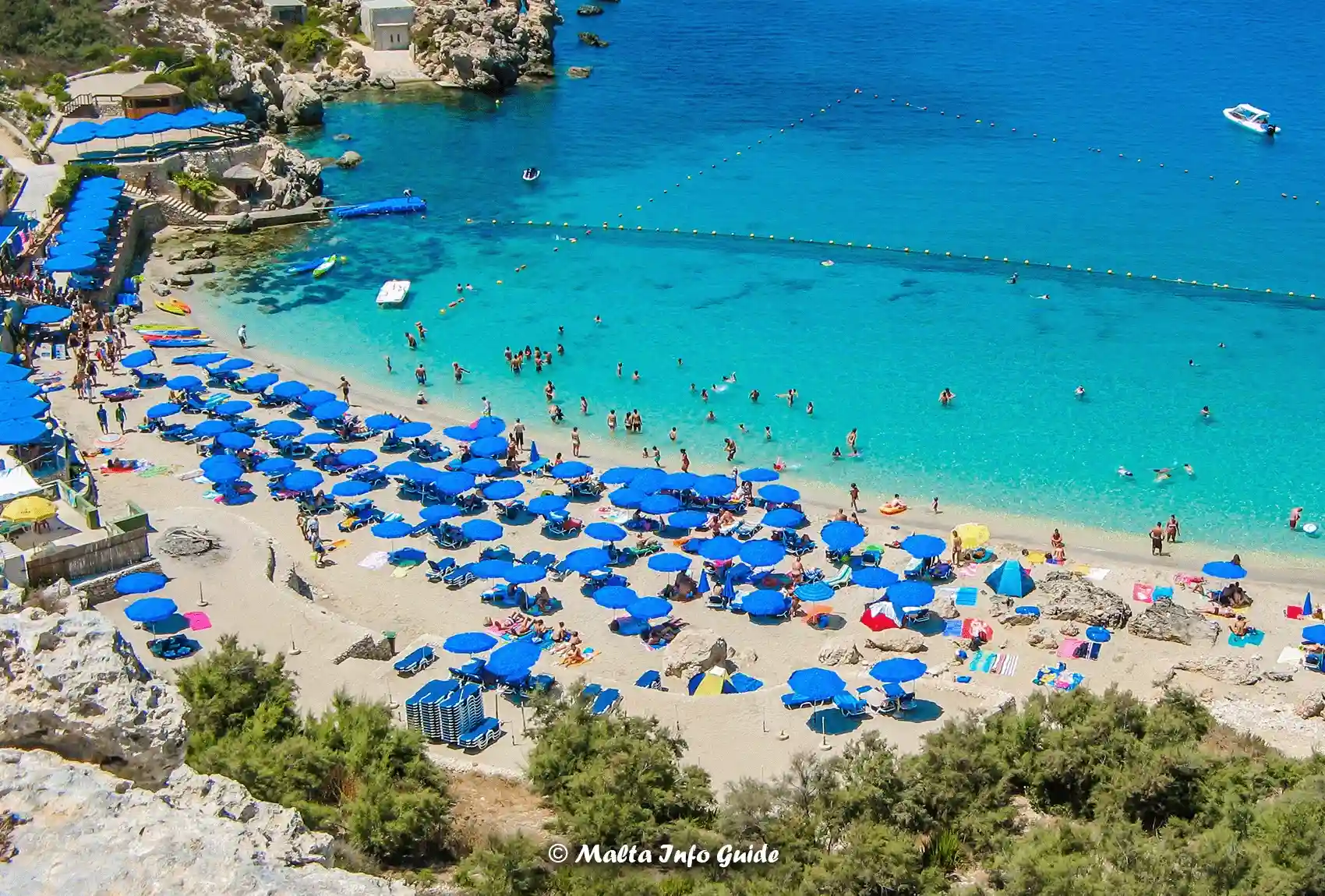 Crowded Paradise Bay beach with blue umbrellas and swimmers.