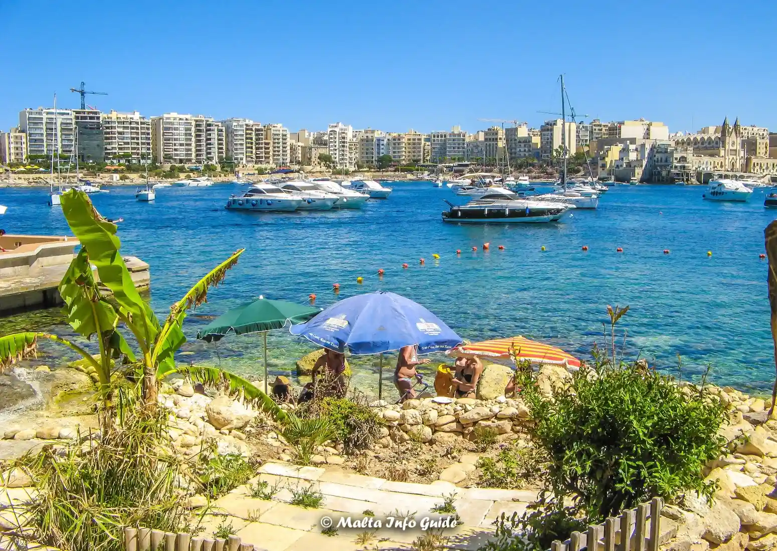 Umbrellas overlook swimmers and boats along Sliema’s rocky shore.