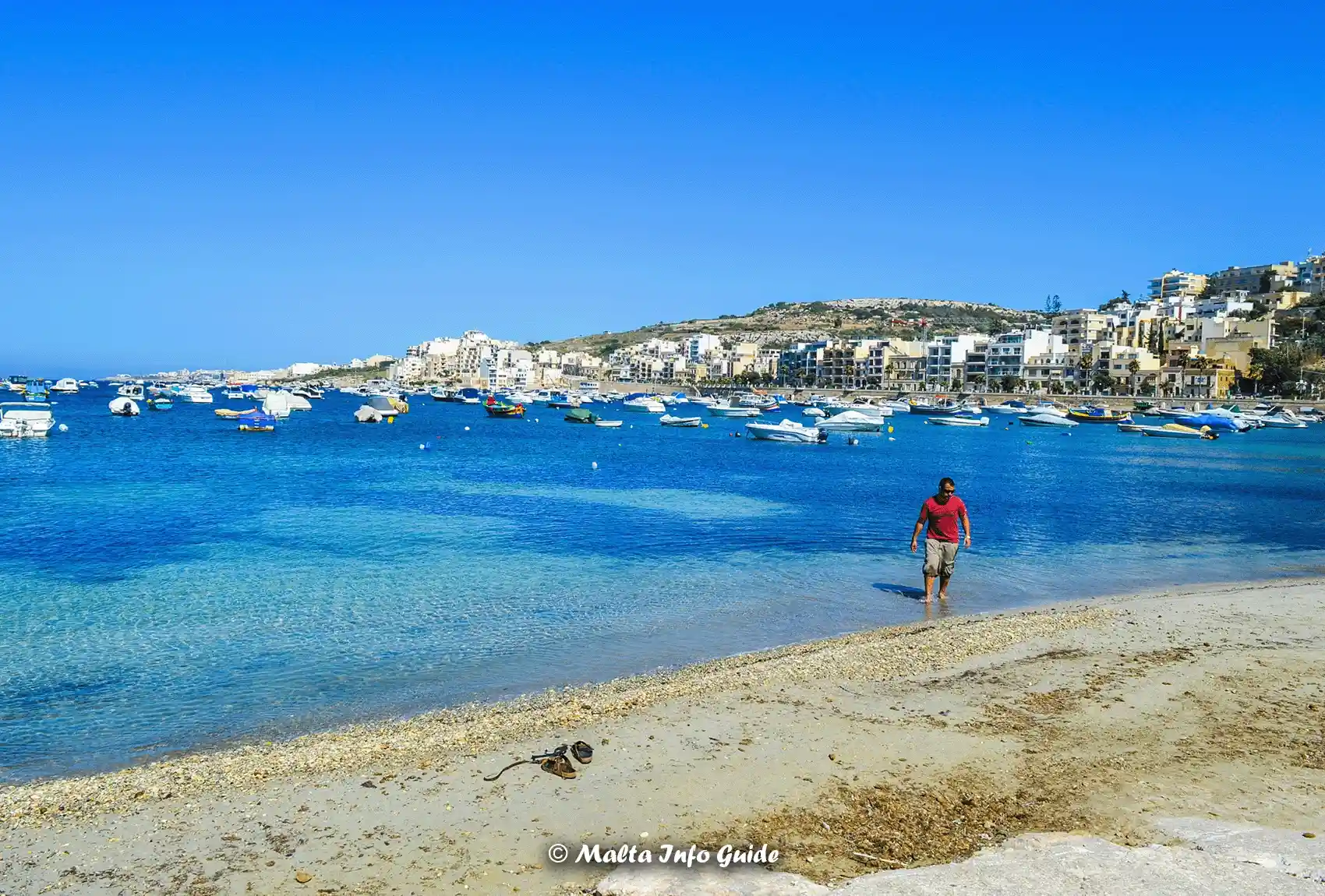 The small sandy beach at Xemxija Bay, with boats on the water and St Paul’s Bay, Buġibba, and Qawra in the background