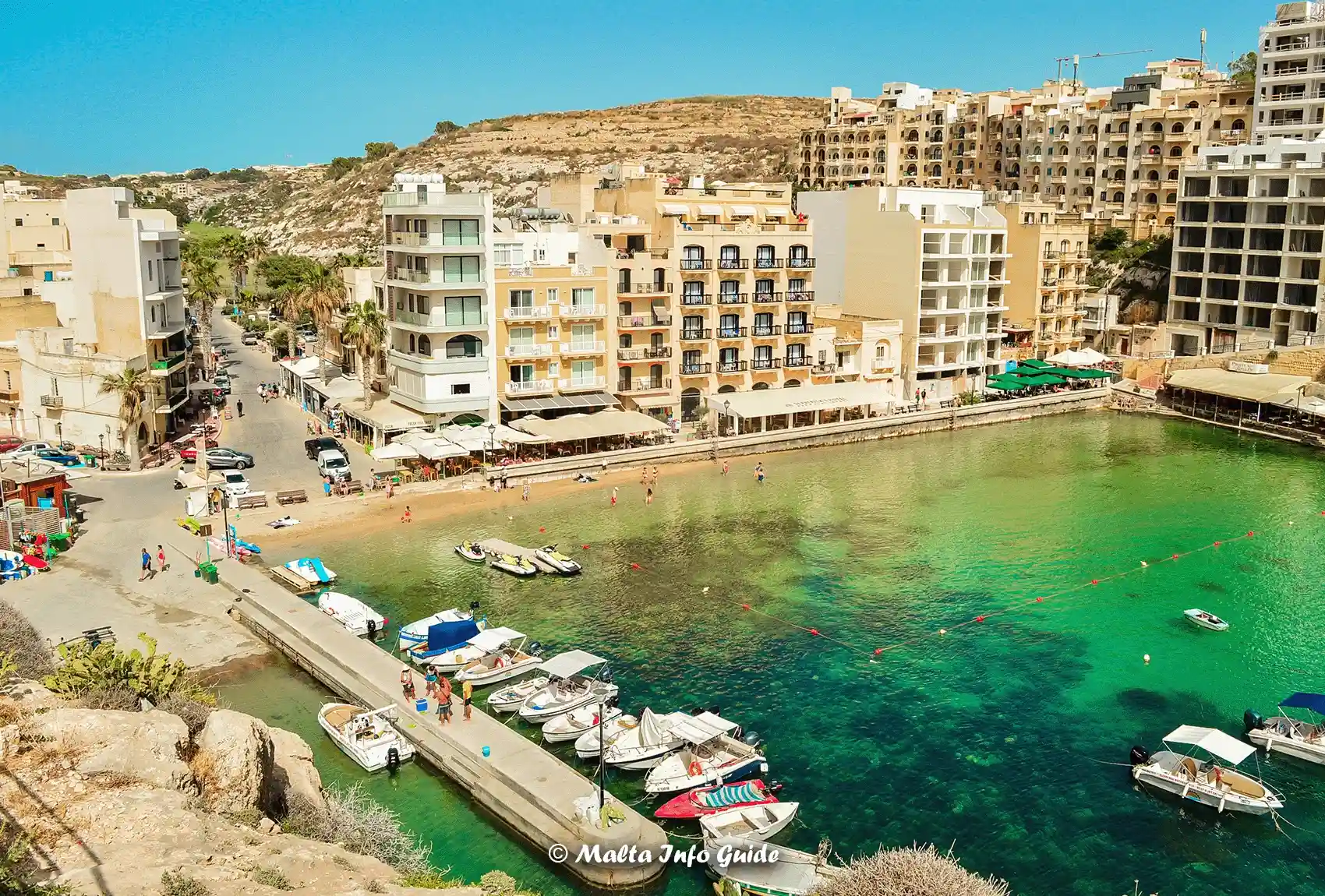Gozo's Xlendi Bay with seafront hotels, restaurants, and boats moored along the jetty.